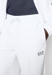 Λευκό παντελόνι φόρμας με ελαστική μέση και κορδόνι, κομψό σχέδιο και μαύρο λογότυπο EA7 στο αριστερό μηρό.