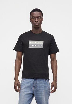 Jeune homme portant un t-shirt noir Jack & Jones et un jean bleu clair, debout devant un fond blanc uni.