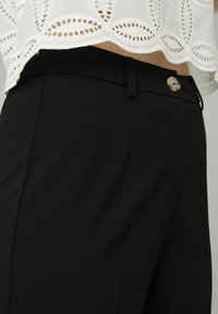 Pantalons noirs ajustés avec une texture lisse, un devant plat et une fermeture à bouton. La taille est soigneusement finie, montrant des détails de couture précis.