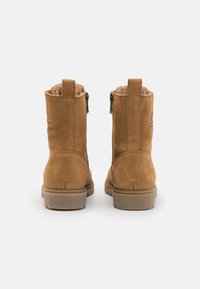 Botas de tornozelo em camurça bege, com forro de pelagem, apresentando um salto baixo e fechos laterais. A parte traseira inclui uma marca gravada no couro.
