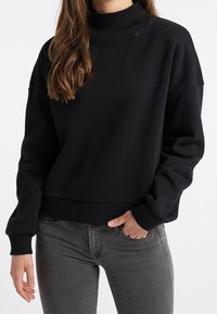 Musta korkeakauluksinen collegepaita, jossa rento istuvuus, resorit hihoissa ja helmassa. Pehmeä kangas, jossa rinnassa hillitty brändäys. Yhdistetty harmaiden farkkujen kanssa.