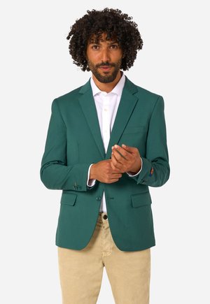 Mann mit lockigem Haar, der einen grünen Blazer über einem weißen Hemd und beigen Hosen trägt, auf einem hellen Hintergrund seinen Blazerärmel zurechtrückt.