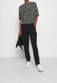 Haut léger à motif vert et blanc, associé à un pantalon cargo noir. Le modèle porte des baskets blanches et tient un sac à main noir.
