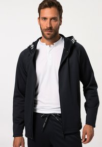 Marineblouson mit Kapuze, ausgestattet mit kontrastierendem weißen Branding auf der Kapuze, getragen über einem weißen Henley-Shirt, mit glatter Textur und sportlichem Schnitt.