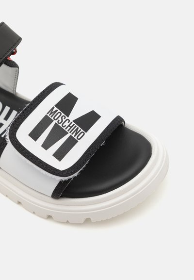 Sandalia deslizante en blanco y negro con tira ajustable, suela de goma y un atrevido logotipo "Moschino" en un diseño geométrico. Acabado mate y ligero.