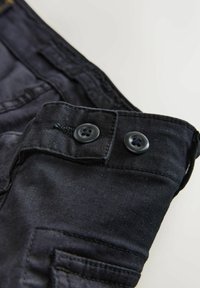 Close-up of black denim pants waistband featuring two black buttons and detailed stitching on a light background.