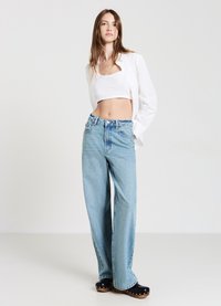 Jeans a gamba larga di colore blu chiaro realizzati in denim, abbinati a un top corto bianco e una camicia bianca aperta, indossati con zoccoli neri.