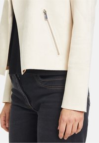 Cream-colored faux suede jacket with a zippered pocket, fitted design, and black textured inner layer layered over dark jeans.