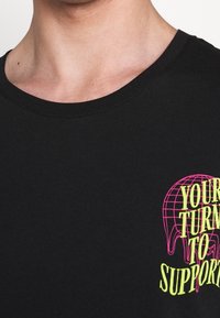 Černé bavlněné tričko s kulatým výstřihem, které na levé straně hrudi zdobí živý neonově růžový a žlutý grafický textový design.