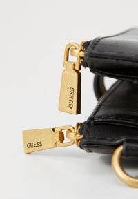 Black leather accessory with gold-engraved zipper pulls marked "GUESS." Features a smooth texture and metal hardware accents.