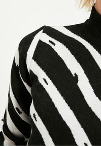 Musta-valkoraidallinen neulepaita korkealla kauluksella. Pehmeä neulottu kangas, jossa on rohkeat, vaakasuorat raidat ja epäsäännöllisiä yksityiskohtia.