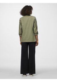 Olivově zelená bunda s ohrnutými rukávy, rovného střihu a detailem na zadním švu. V kombinaci s kalhotami širokého střihu černé barvy a plátěnými botami.