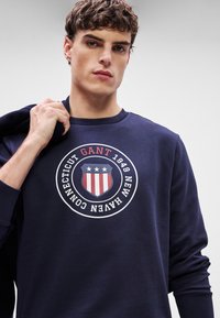 Granatowy sweatshirt z okrągłym logo w czerwonych i białych akcentach, z napisem "GANT", rokiem "1949" oraz napisem "NEW HAVEN CONNECTICUT". Miękka tkanina.