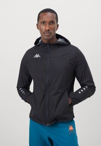Sort zip-up jakke med en tætsiddende design, der har hætte, lys grå indvendig og Kappa-logo accenter på brystet og ærmerne.