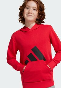 Roter Hoodie mit einem schwarzen dreieckigen Grafikdesign auf der Vorderseite, Kängurutasche und verstellbarem Kordelzug für die Kapuze. Weicher Stoff und gerippte Bündchen.