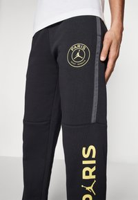 Czarne spodnie dresowe z żółtym logo Paris Saint-Germain i napisem "PARIS". Szary pas boczny i miękka, teksturowana tkanina.