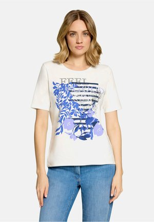 Femme aux cheveux blonds mi-longs portant un t-shirt blanc avec un motif graphique bleu à fleurs et rayures et un jean bleu.