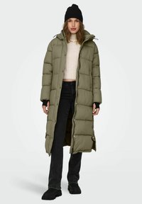 Olivegrüner langer Daunenmantel mit Kapuze, breiten quilted Paneelen, Druckknopfverschlüssen und Seitentaschen, kombiniert mit schwarzen Jeans und einer Mütze.