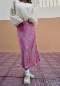 Woman wearing a white fuzzy sweater, pink skirt with green polka dots, white socks, and beige slip-on shoes standing on tiled floor.