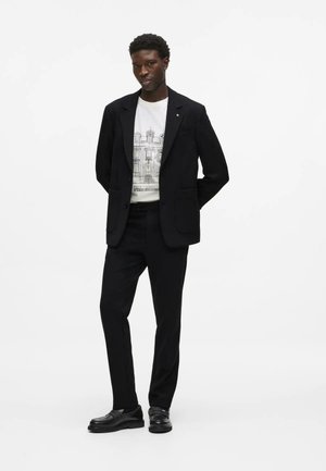 Muž stojaci so založenými rukami, oblečený v čiernom obleku, biele tričko s grafikou a čierne mokasíny na bielom pozadí.