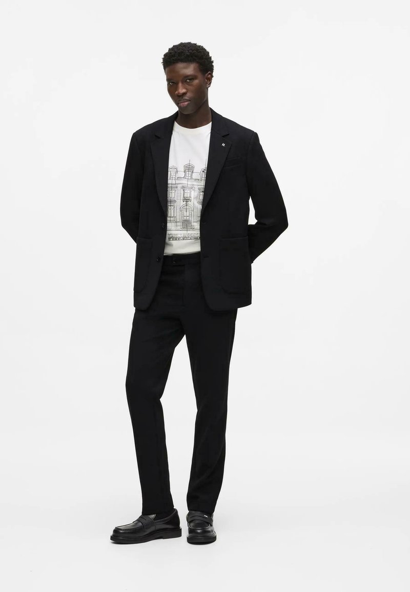 Homme debout, mains derrière le dos, portant un costume noir, un t-shirt blanc avec un motif graphique et des mocassins noirs sur fond blanc.