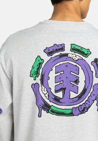 Lila und grün stilisierte kreisförmige Grafik mit abstrakten Formen und strukturierten Details auf der Rückseite eines hellgrauen Sweatshirts.
