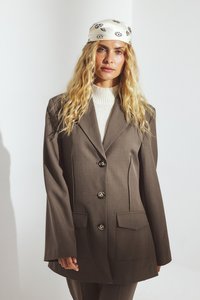 Blazer marrone su misura con tre grandi bottoni, due tasche frontali e rever larghi. Abbinato a un maglione bianco a coste sotto.