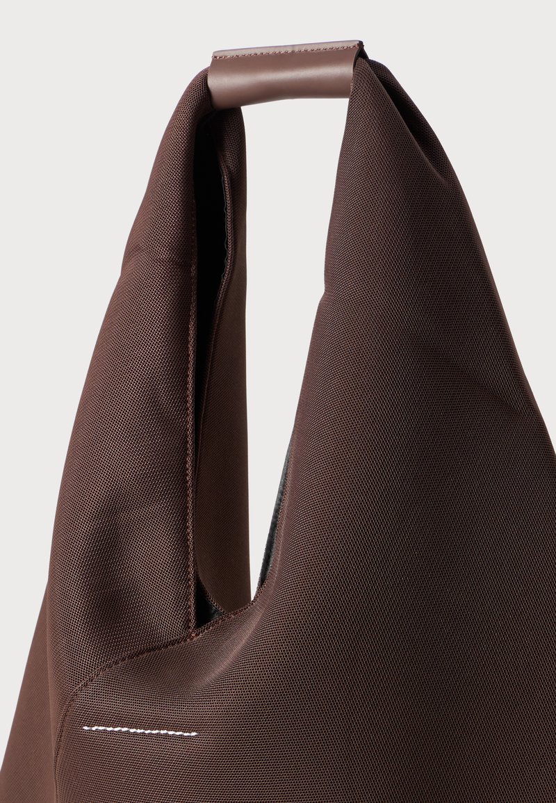 Brown tote bag made of textured fabric, featuring a unique handle design and a visible white stitching detail.
