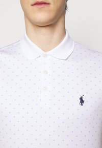Witte polo met een gestructureerd patroon van kleine blauwe diamanten, drie knopen en een donkerblauw geborduurd logo op de linkerkant van de borst.