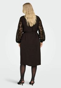 Czarna sukienka z długimi, przezroczystymi koronkowymi rękawami, paskiem w talii oraz o długości do kolan. Materiał wydaje się mieć fakturę i jest elegancki.