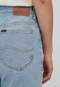 Světle modré džíny z denimu s koženou nášivkou na pasu, žlutým prošíváním a zadní kapsou s černou značkovou visačkou.