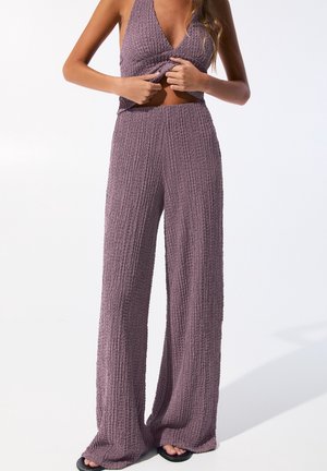 Femme ajustant un haut sans manches texturé lavande associé à un pantalon large assorti et des sandales noires sur fond blanc.