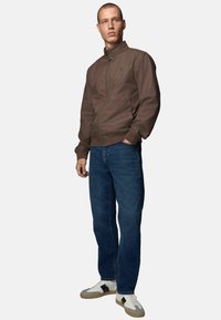 Veste légère en brun à fermeture éclair avec col montant, associée à un jeans en denim bleu et des baskets blanches avec des accents gris et des semelles texturées.