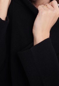 Czarny dziany sweter z prążkowanymi mankietami, z wysokim kołnierzem podciągniętym do góry. Ręka z złotym pierścieniem trzymająca krawędź kołnierza. Miękka faktura.