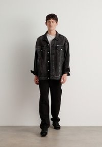 Chaqueta de mezclilla negra con dos bolsillos en el pecho, cierre de botones y corte relajado, combinada con pantalones negros y zapatos oscuros, sobre un fondo neutro.