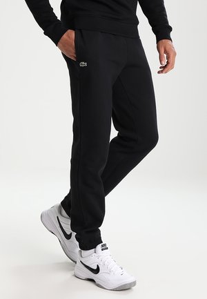 Uomo che indossa pantaloni sportivi neri Lacoste e scarpe da ginnastica bianche Nike, in piedi con una mano nella tasca su sfondo chiaro.