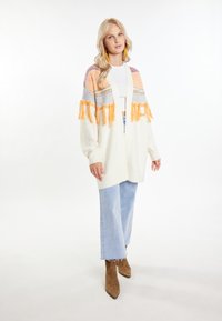 Krämfärgad cardigan med flerfärgade stickade mönster och fransar i orange, grått och rosa; kombinerad med ljusblå vida jeans och bruna klackstövlar.