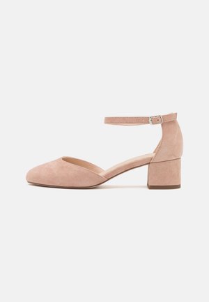 Beige nupuk mid-korkokenkä, jossa on suljettu pyöreä varvas, säädettävä ankle-strap ja lohkokorko valkoista taustaa vasten.
