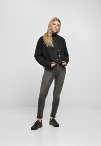 Černý pletený cardigan s velkými knoflíky, kombinovaný s černým topem a šedými slim kalhotami. Model má na sobě černé tenisky. Texturovaná tkanina.