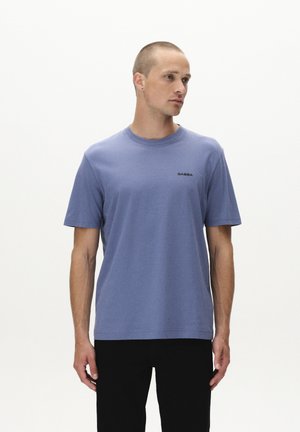 Blue cotton t-shirt with a crew neck and short sleeves. Features a small black logo on the chest, paired with dark pants.