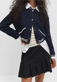 Marineblau gestrickte Strickjacke mit weißem Saum, Knopfverschluss und Taschen, getragen über einem weißen Hemd und kombiniert mit einem schwarzen, plissierten Rock.