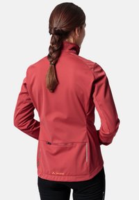 Rød softshell-jakke med høy krage, med en glidelåslomme bak, kontraststing og logo nederst. Glatt tekstur.