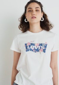 Balta medvilnė marlės marškinėliai su mėlyna gėlių Levi's logotipo grafika priekyje. Trumpomis rankovėmis ir laisvu siluetu.