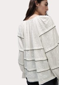 Blusa bianca con volant sovrapposti bordati in nero, tessuto strutturato e scollatura ampia. Le maniche sono larghe e fluenti.