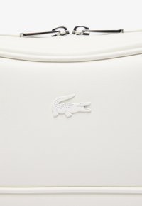 Borsa bianca dalla texture con un logo di coccodrillo bianco centrato sotto due zip argentate su uno sfondo chiaro.