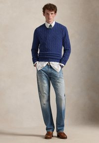 Jeune homme aux cheveux bouclés portant un pull bleu à torsades par-dessus une chemise blanche avec cravate, un jean déchiré et des mocassins marron, debout les mains dans les poches.