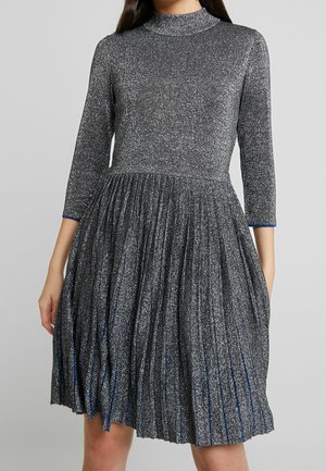 Frau trägt ein langärmeliges, hochgeschlossenes, dunkelgraues glitzerndes Kleid mit einem plissierten Rock, der über den Knien endet, vor unifarbenem Hintergrund.