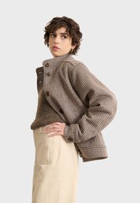 Brown and blue checked jacket with a high collar, button closure, and oversized sleeves, paired with a textured brown sweater and beige pants.