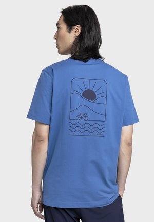 Mann mit schulterlangem Haar, der ein blaues T-Shirt trägt, das auf der Rückseite eine schwarze Grafik mit Sonne, Hügeln, Fahrrad und Wellen zeigt.