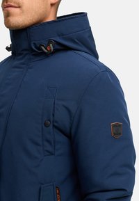 Hombre con una chaqueta azul oscura de alto cuello y toggles ajustables, que cuenta con un bolsillo en el pecho y un parche con el logo en la manga.